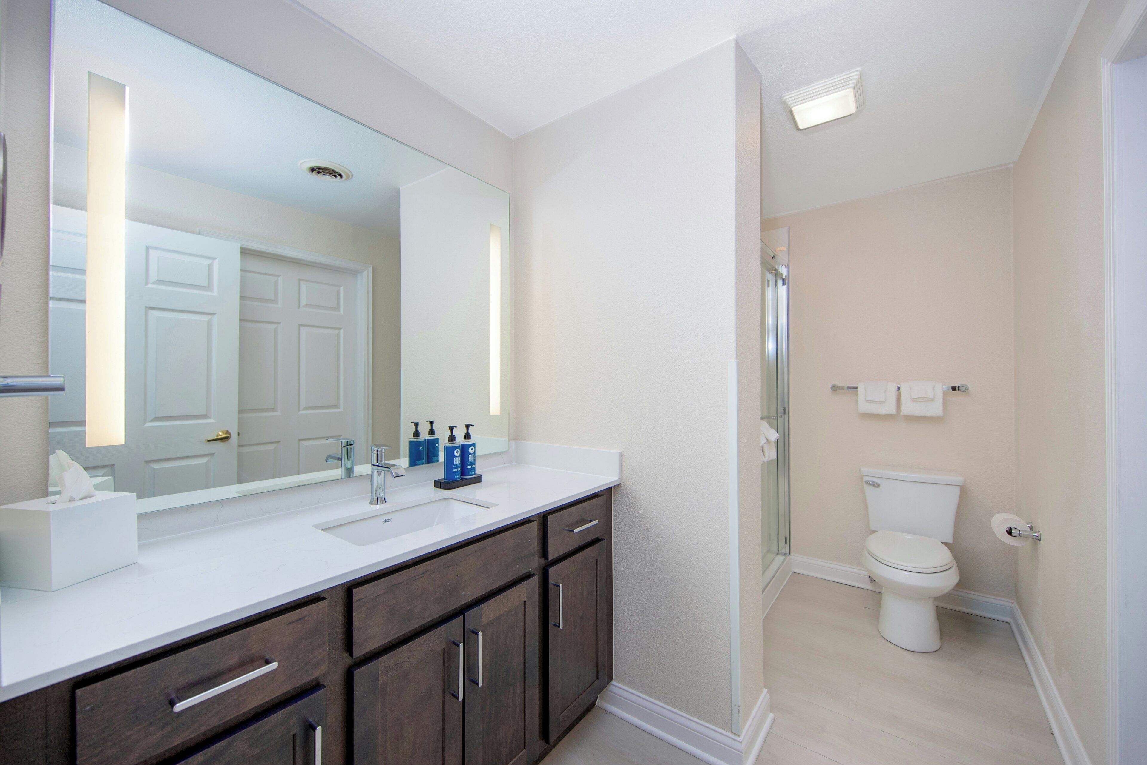 Une salle de bain avec un lavabo blanc, des meubles en bois foncé, un grand miroir, des toilettes et un espace douche.