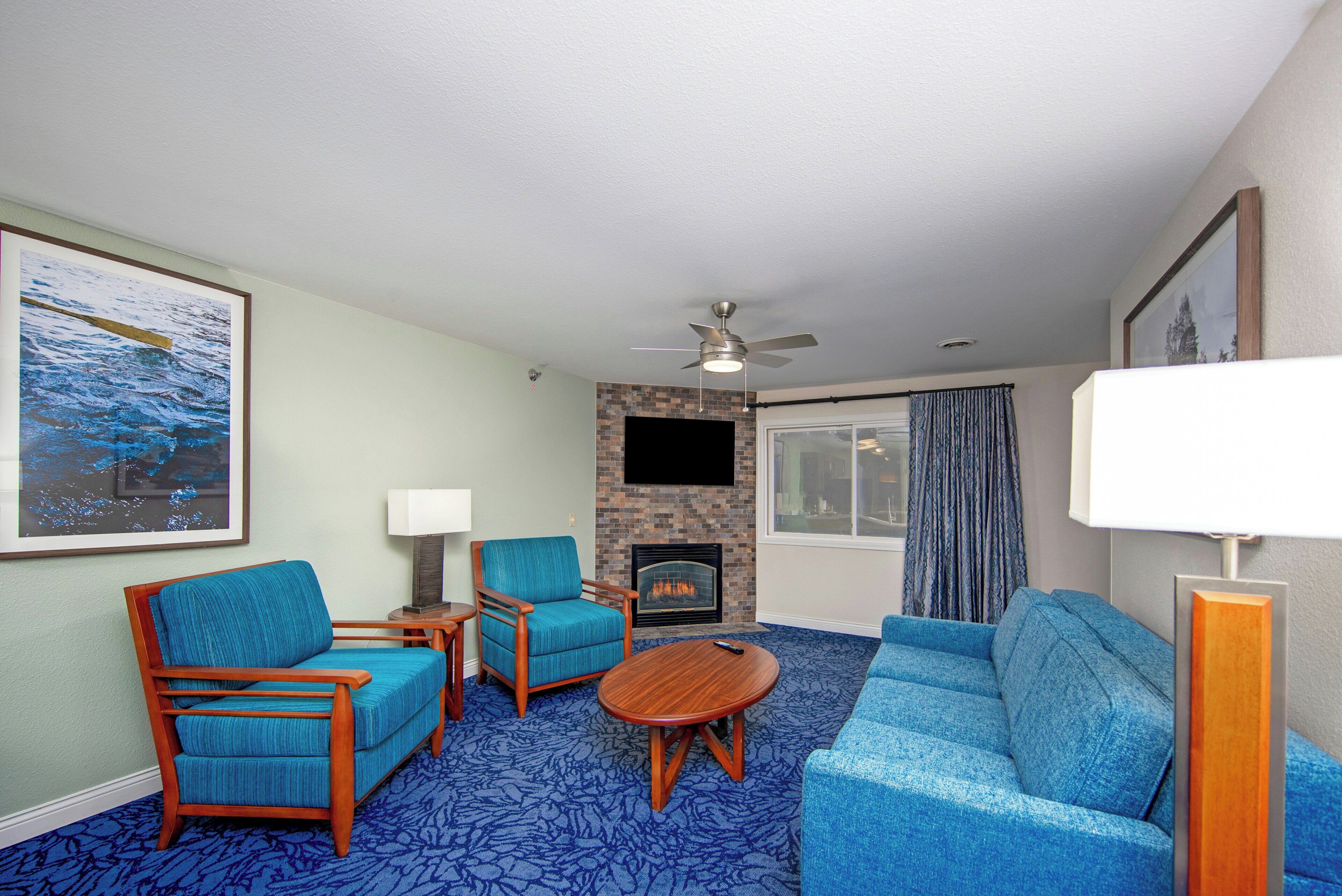 Une chambre d’hôtel avec une cheminée, un ventilateur de plafond, une grande fenêtre avec des rideaux et une moquette bleue.