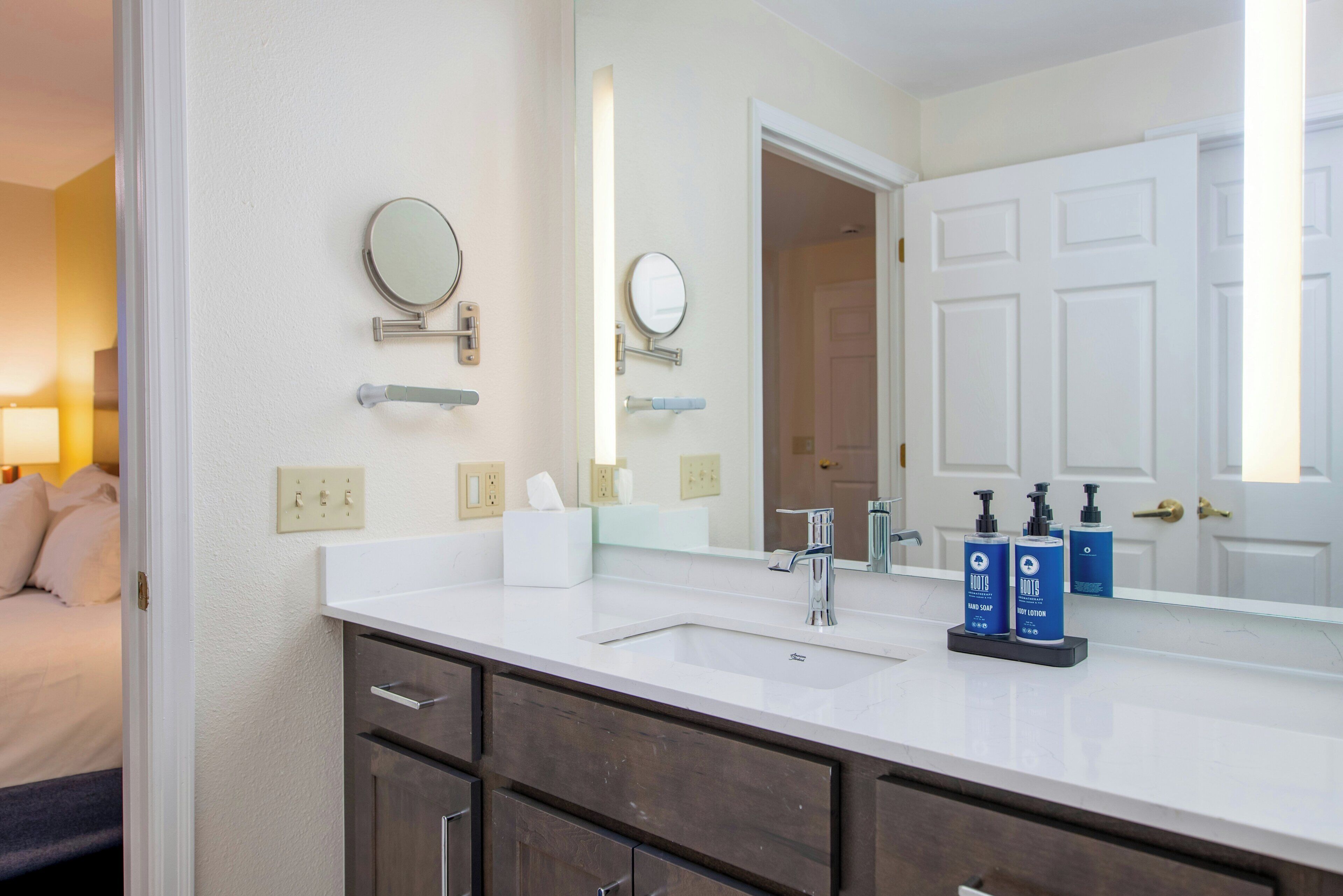 A bathroom with a white sink, two mirrors, a towel dispenser, and a door leading to another room.