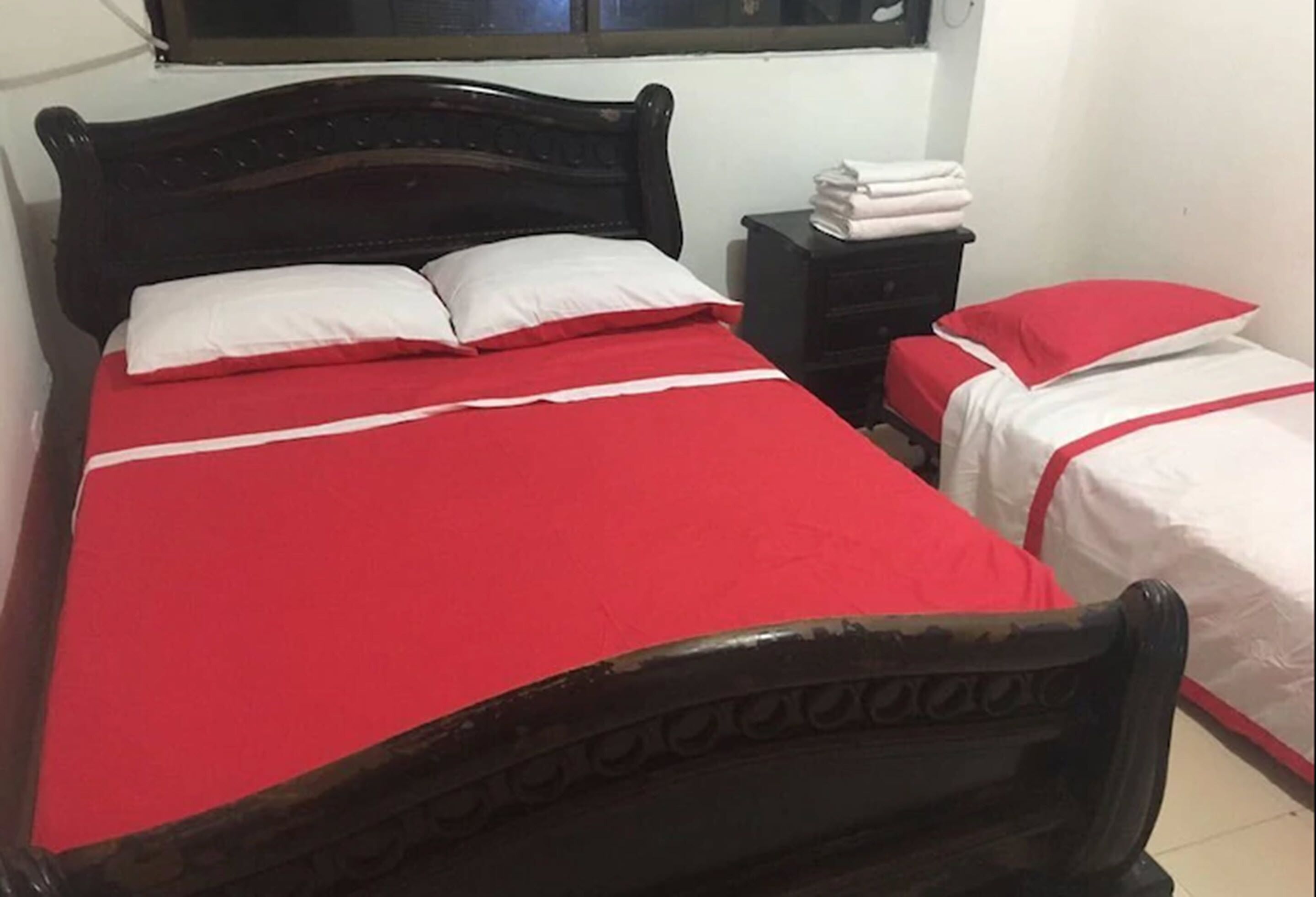 Una habitación con dos camas, cada una con colcha roja y almohadas blancas. Hay una mesita de noche con toallas dobladas y una pequeña mesa junto a las camas.