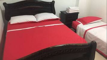 Una habitación con dos camas, cada una con colcha roja y almohadas blancas. Hay una mesita de noche con toallas dobladas y una pequeña mesa junto a las camas.