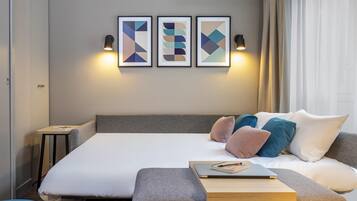 A modern hotel room with a large bed, a small side table, a bedside lamp, and three abstract wall art pieces.