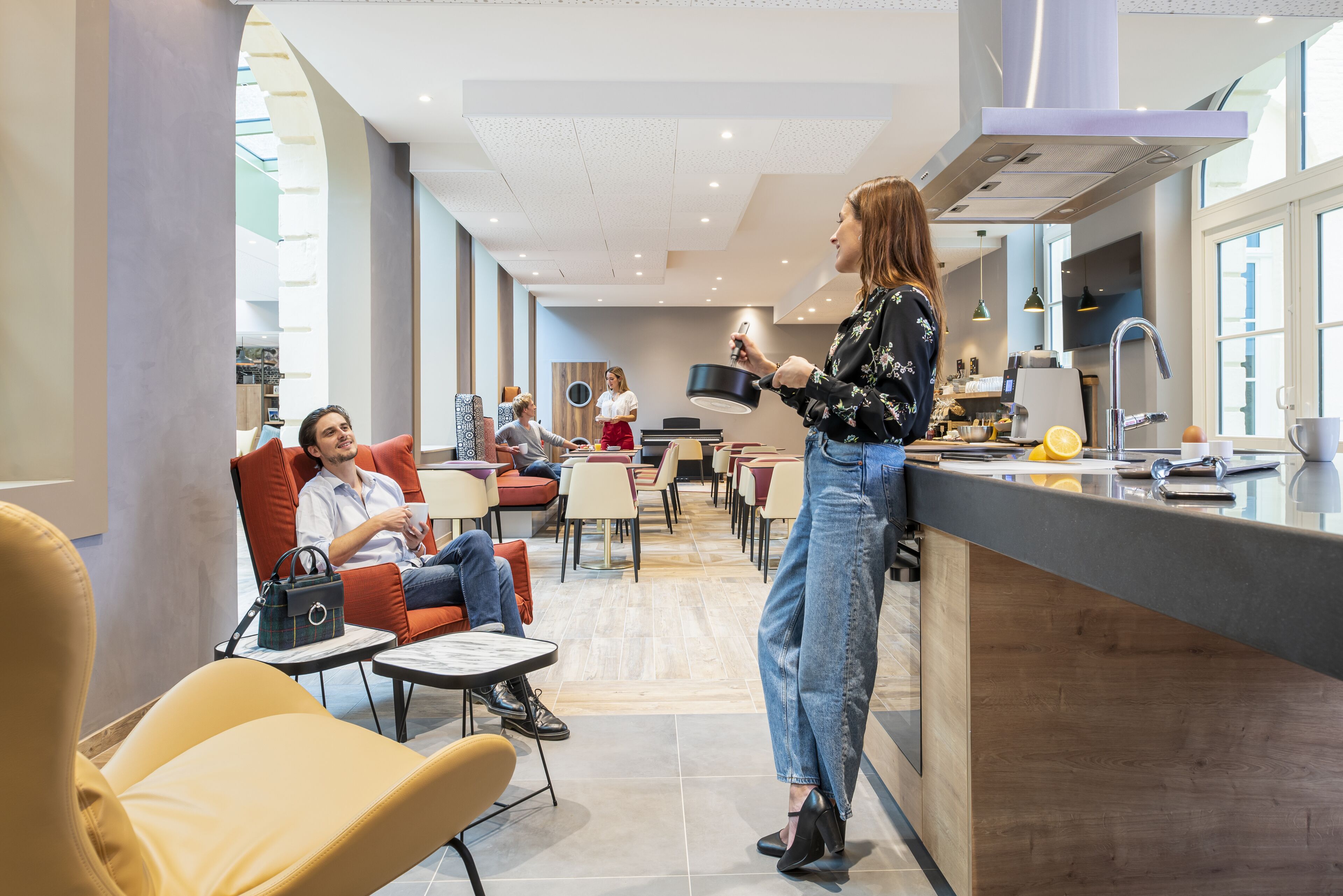 A modern hotel lobby with a bar area, seating arrangements, and a visible kitchen.