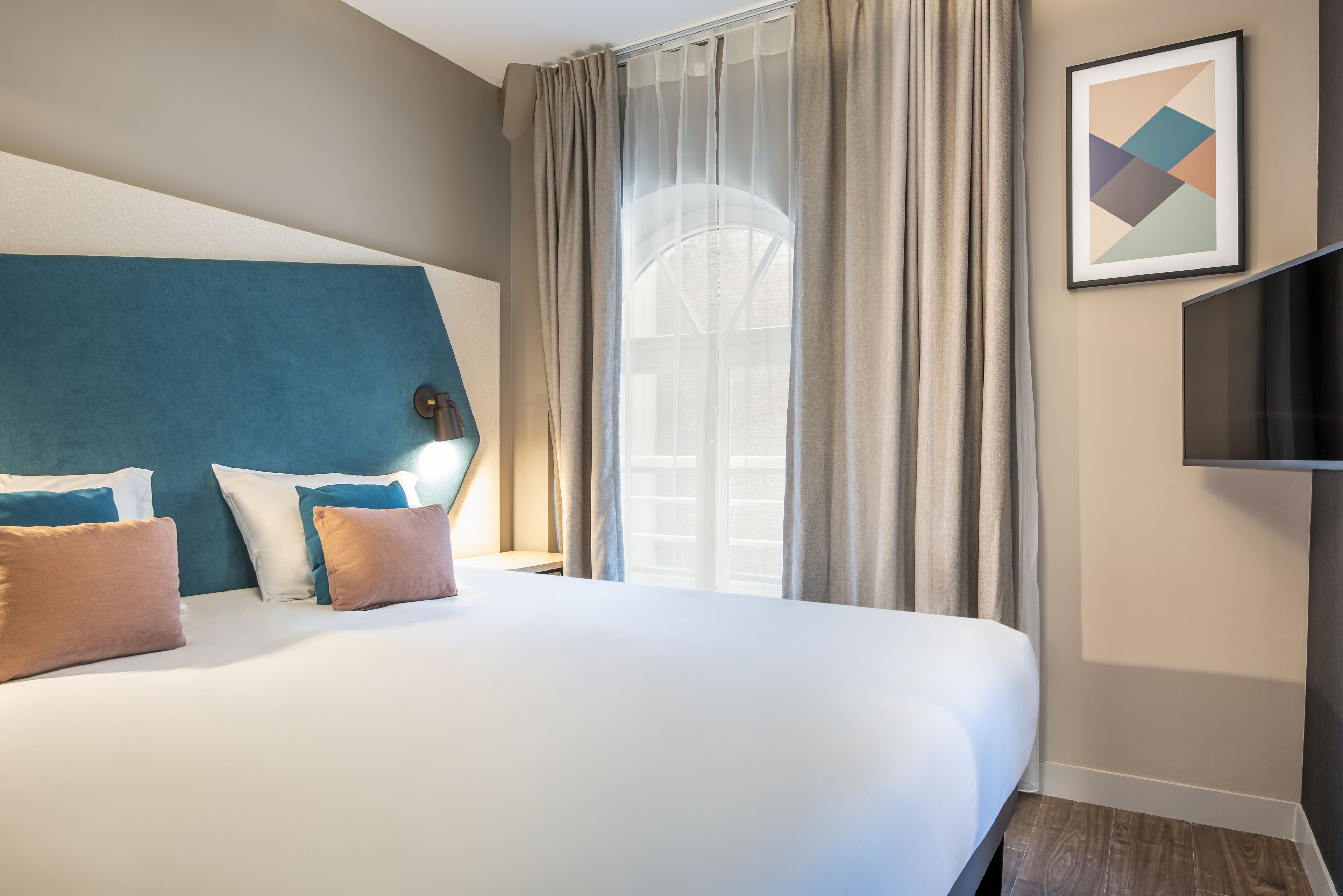 A hotel room with a large bed, a headboard, a TV, and a window with curtains.