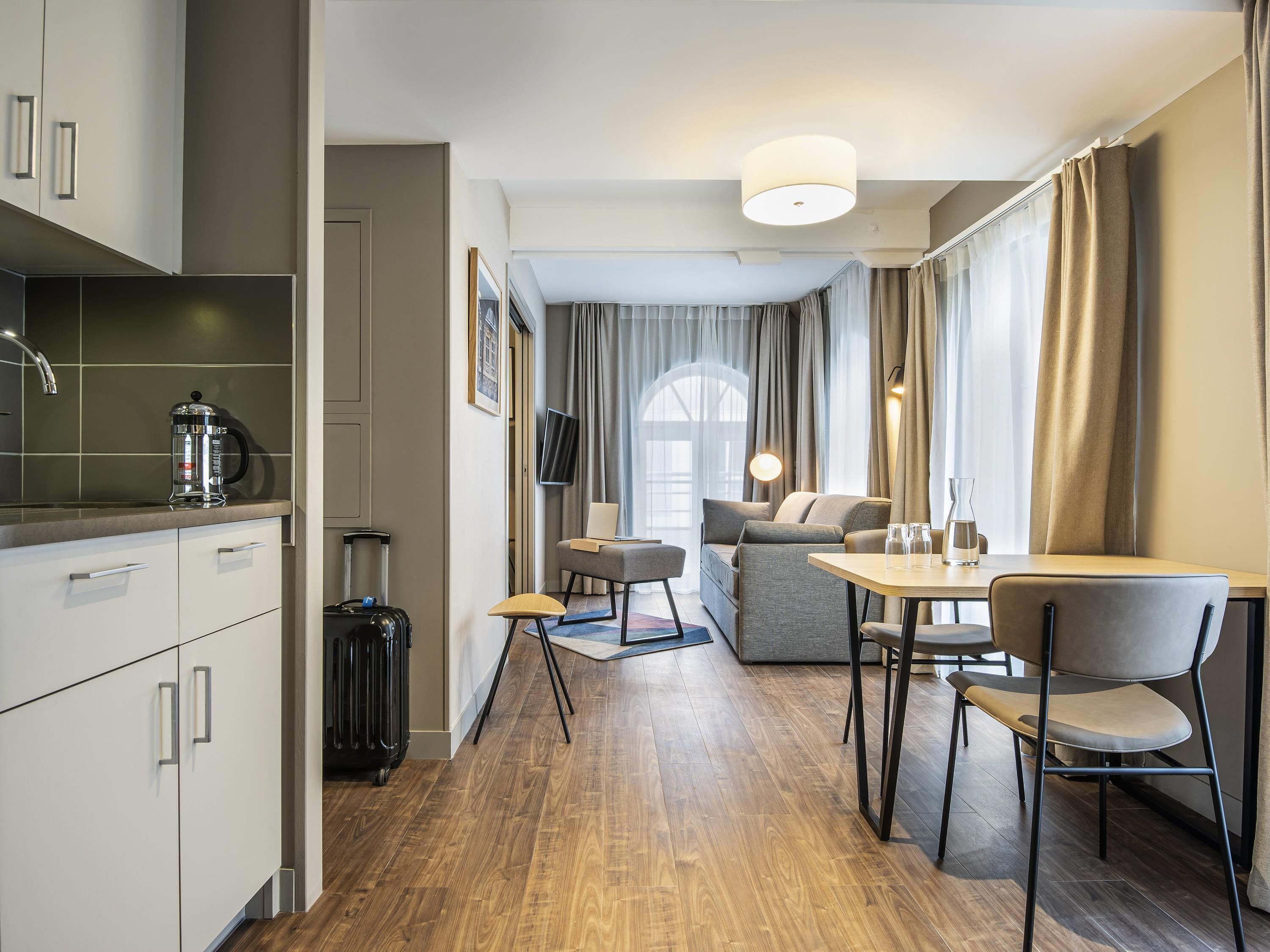 Una habitación de hotel moderna con kitchenette, comedor y zona de estar con sofá y sillones.