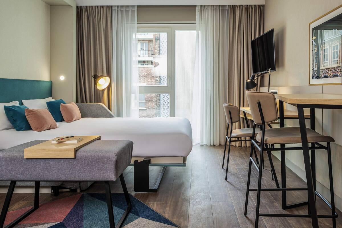 Ein Hotelzimmer mit Bett, Schreibtisch, Stuhl, Fernseher und einem Fenster mit Vorhängen.