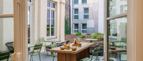 An outdoor dining area with a wooden table and green chairs, surrounded by buildings.