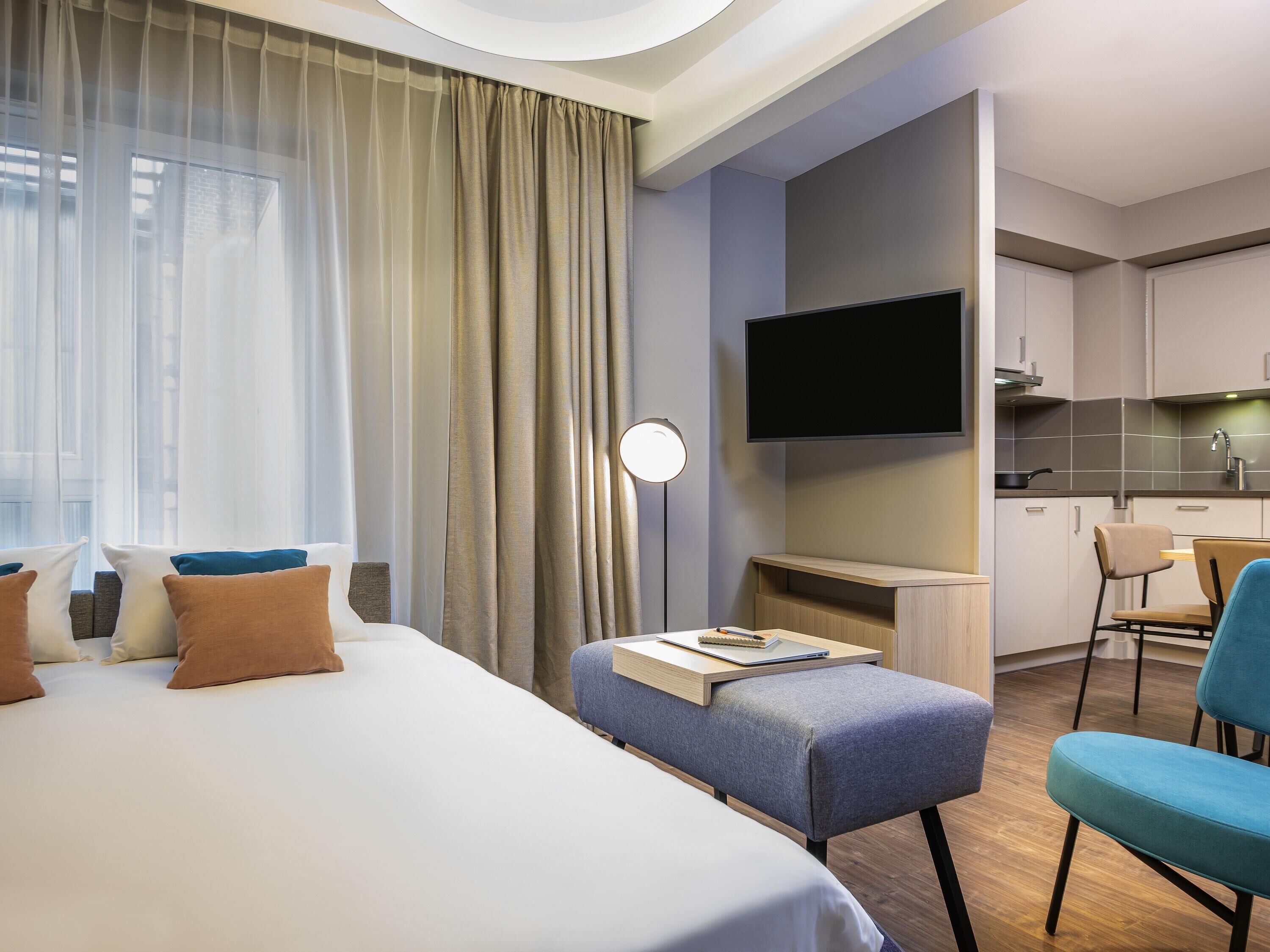 A modern hotel room with a bed, a desk with a chair, a TV, and a kitchenette.