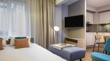 A modern hotel room with a bed, a desk with a chair, a TV, and a kitchenette.