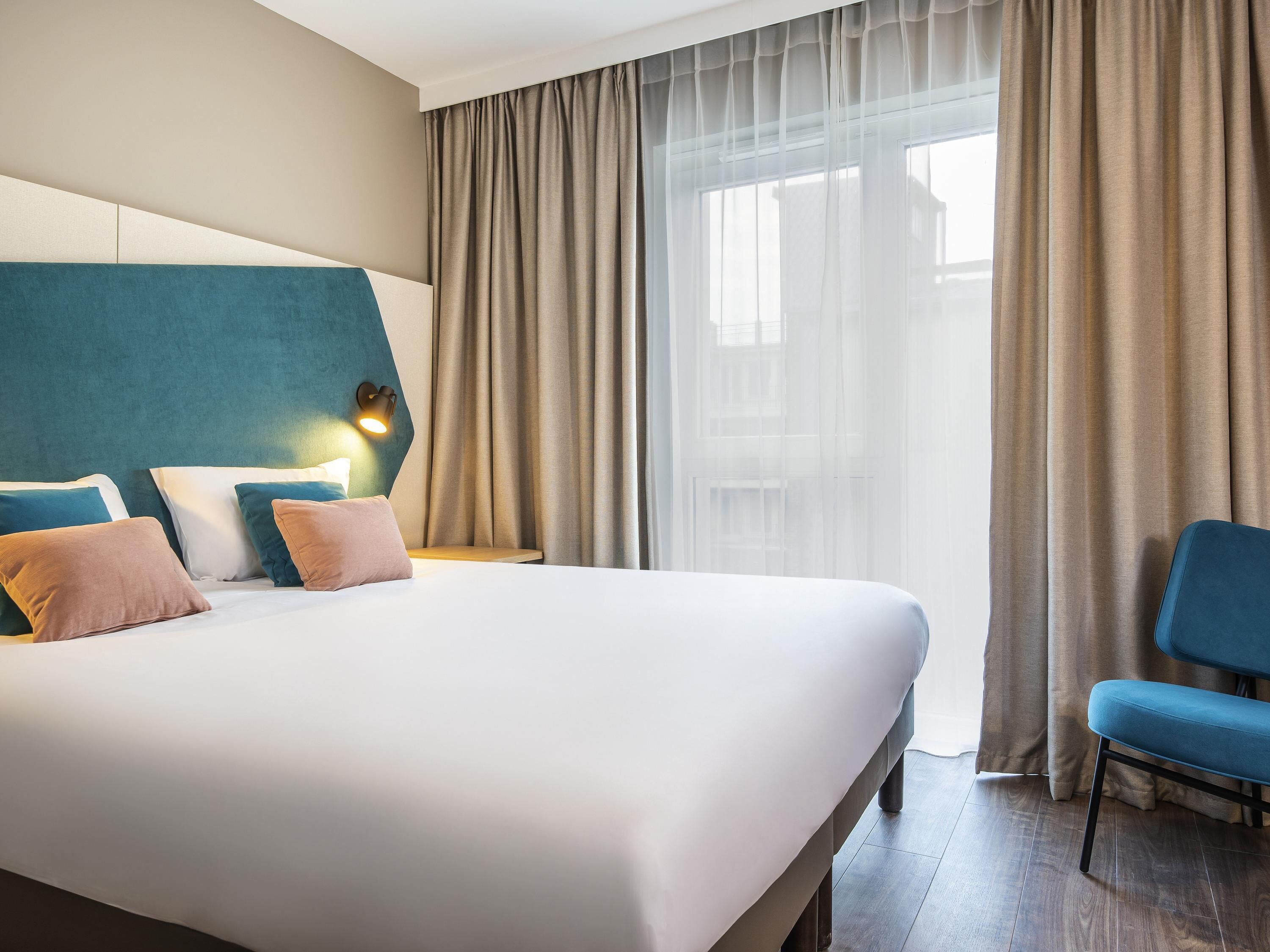 Een hotelkamer met een groot bed, een blauwe stoel en uitzicht op de stad door dunne gordijnen.