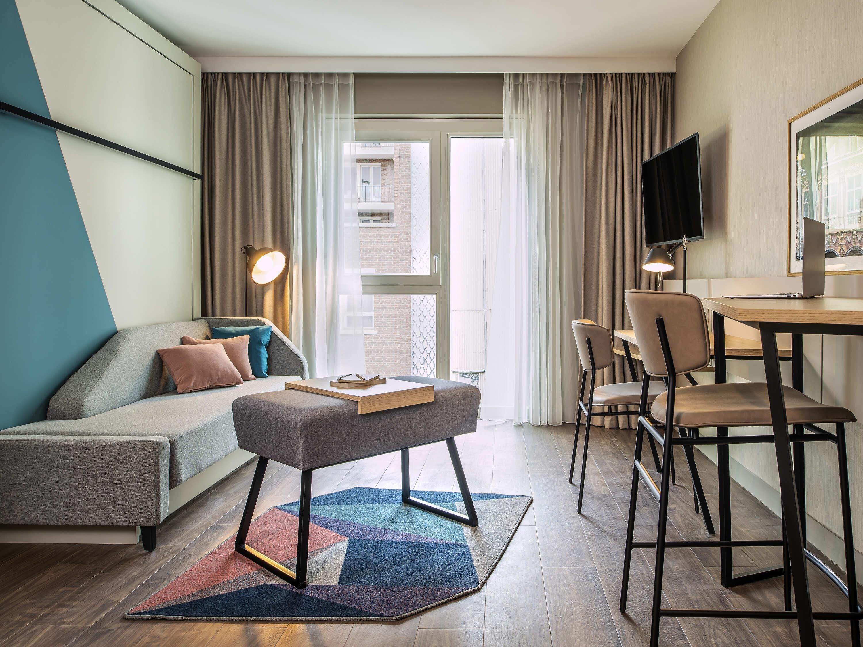 A modern hotel room with a sofa, a small table, a desk with a chair, a TV, and a view of the outside.