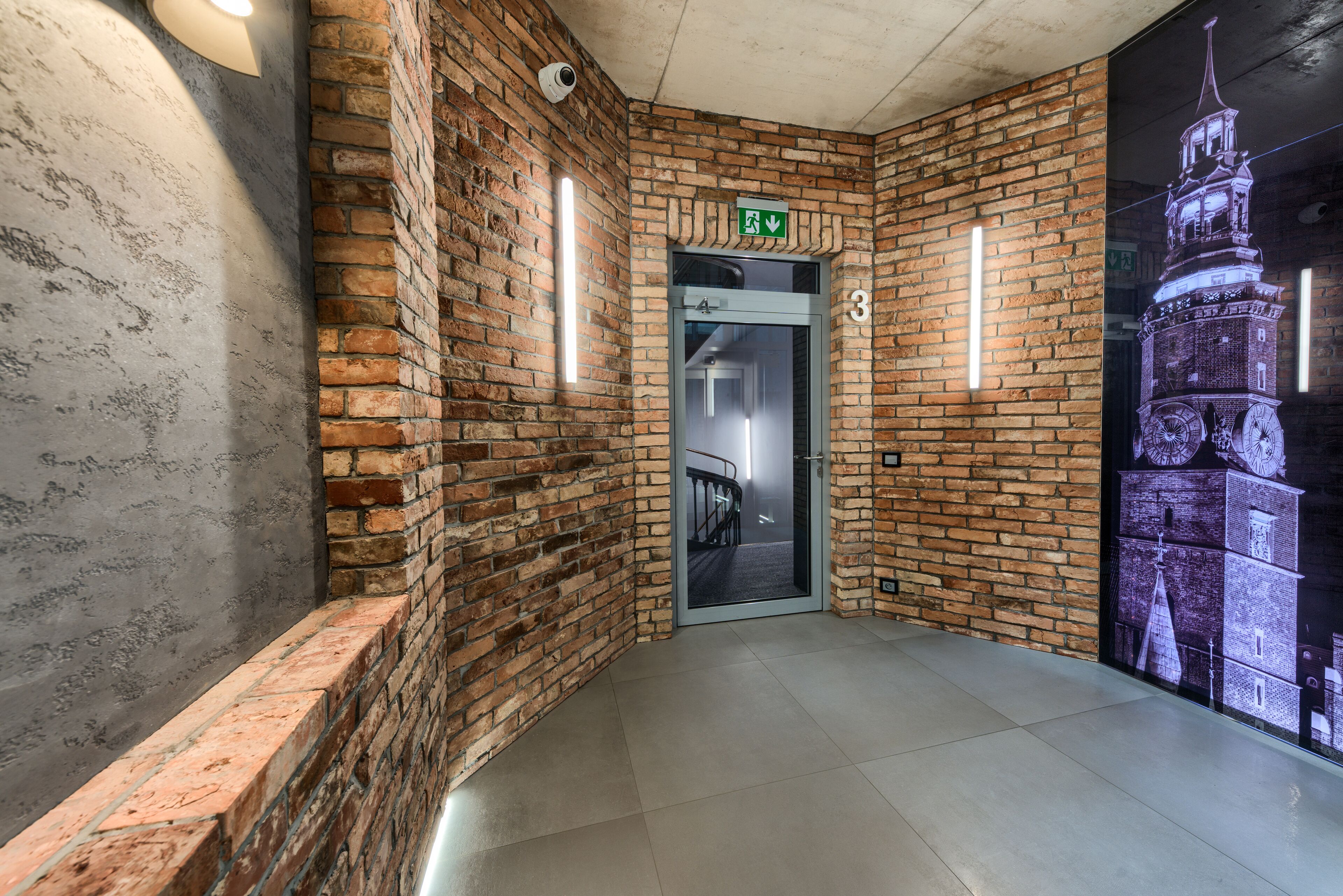 A hallway with brick walls, a glass door marked with an exit sign, and a large illuminated clock tower mural.