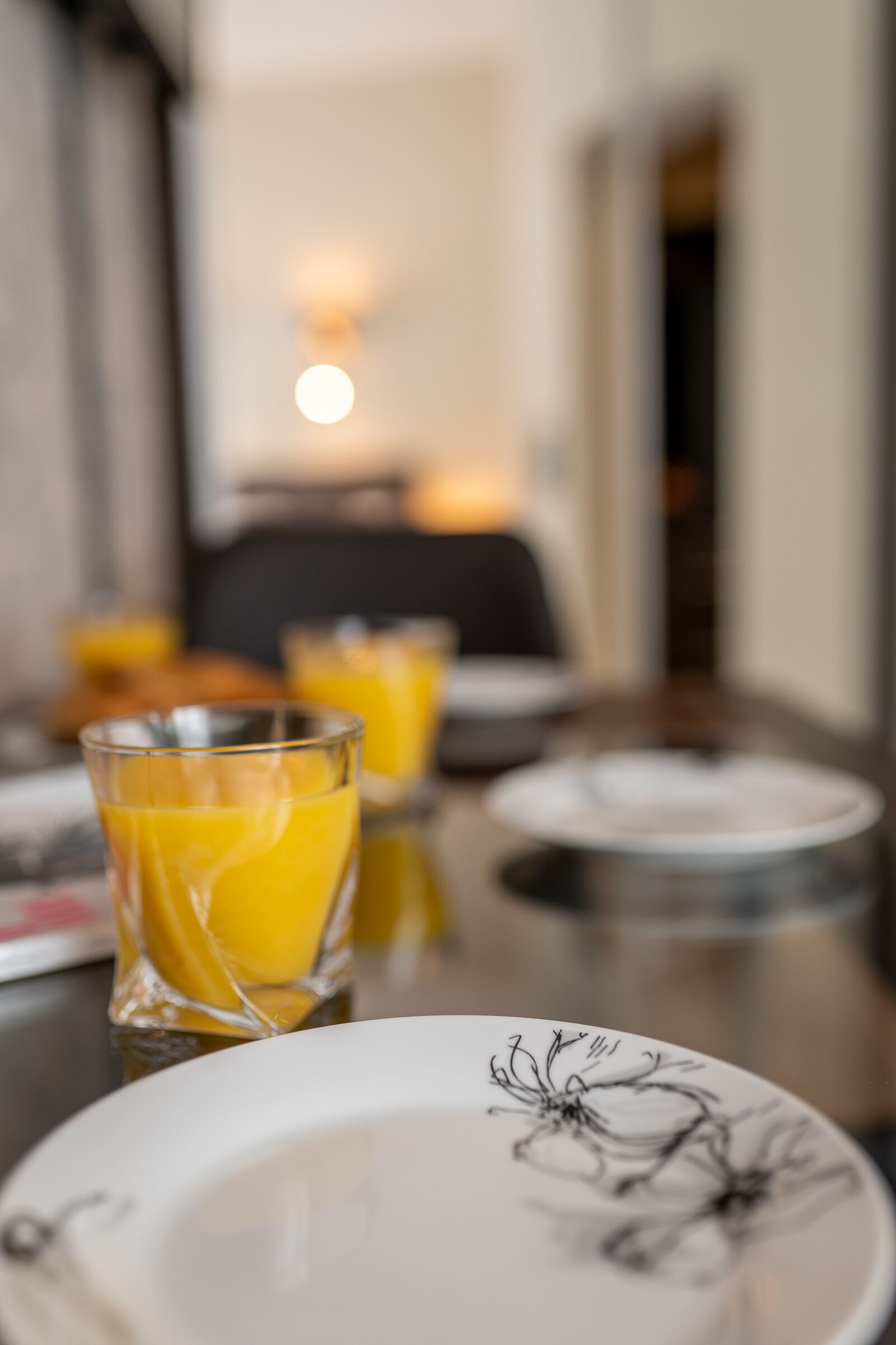 A breakfast table with a glass of orange juice, a plate with a floral design, and a blurred background suggesting a dining area.