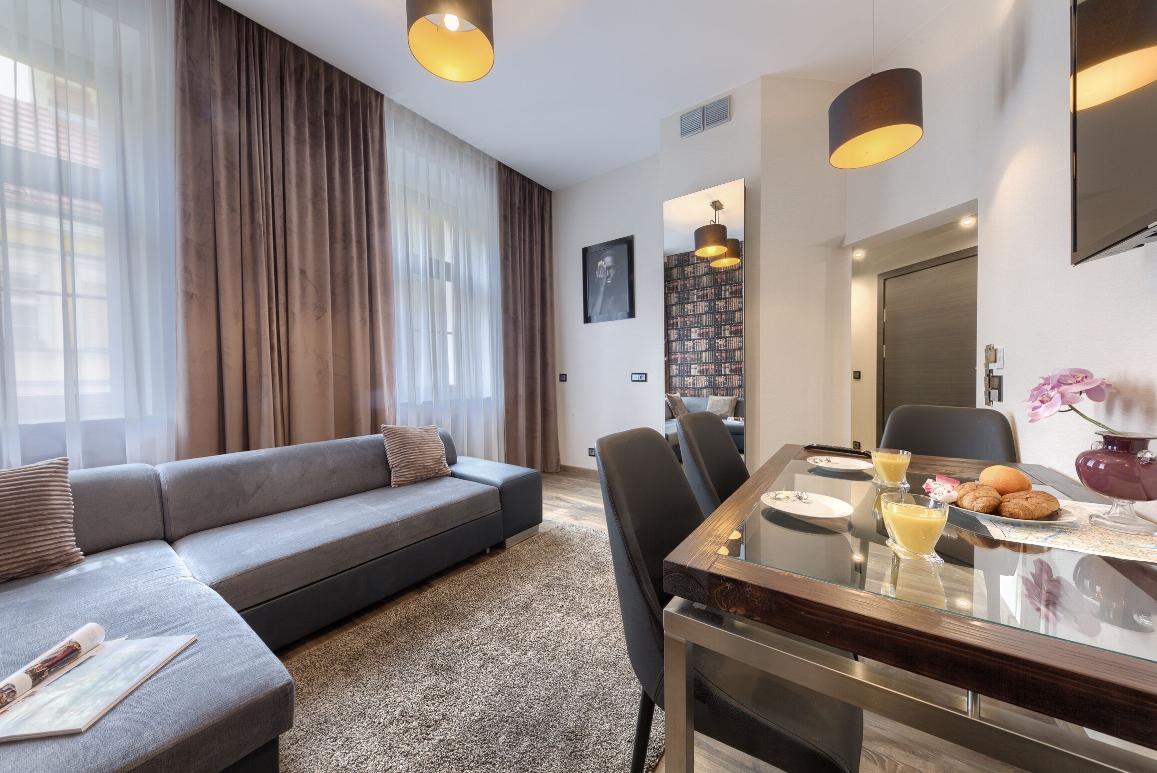 A modern living room with a grey sofa, a dining table with chairs, and a large window with curtains.