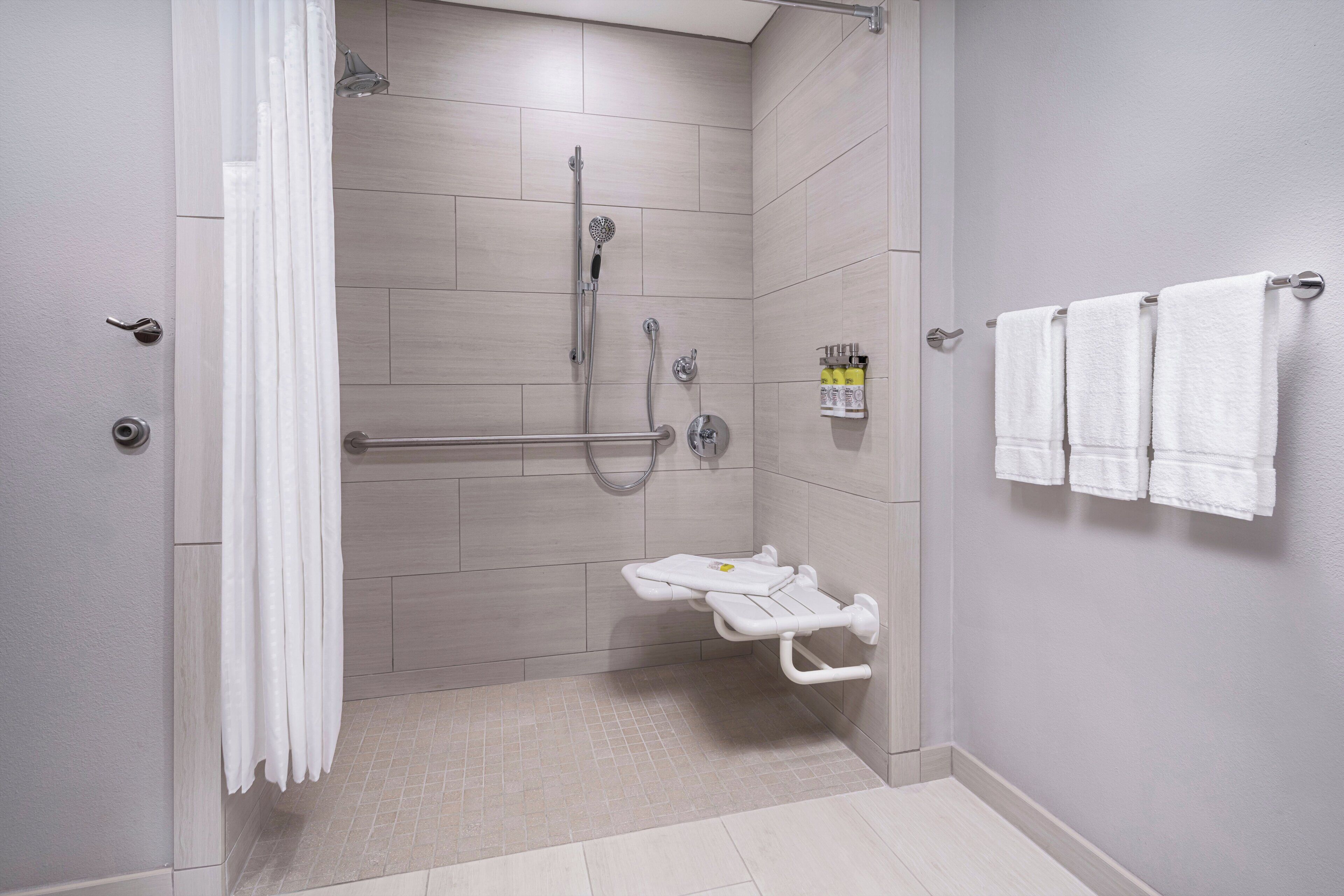 Une salle de bain moderne dotée d’une douche à l’italienne, de barres d’appui, de toilettes avec un siège de sécurité et d’un porte-serviettes muni de plusieurs serviettes.