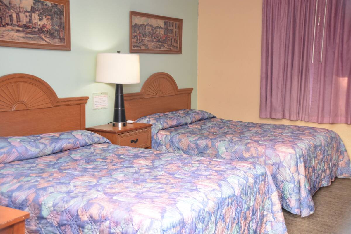 A hotel room with two beds, a nightstand, a lamp, and two framed pictures on the wall.
