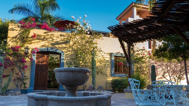 Un patio con una fuente de piedra, sillas de hierro forjado blanco y un edificio con puerta de madera y balcón.