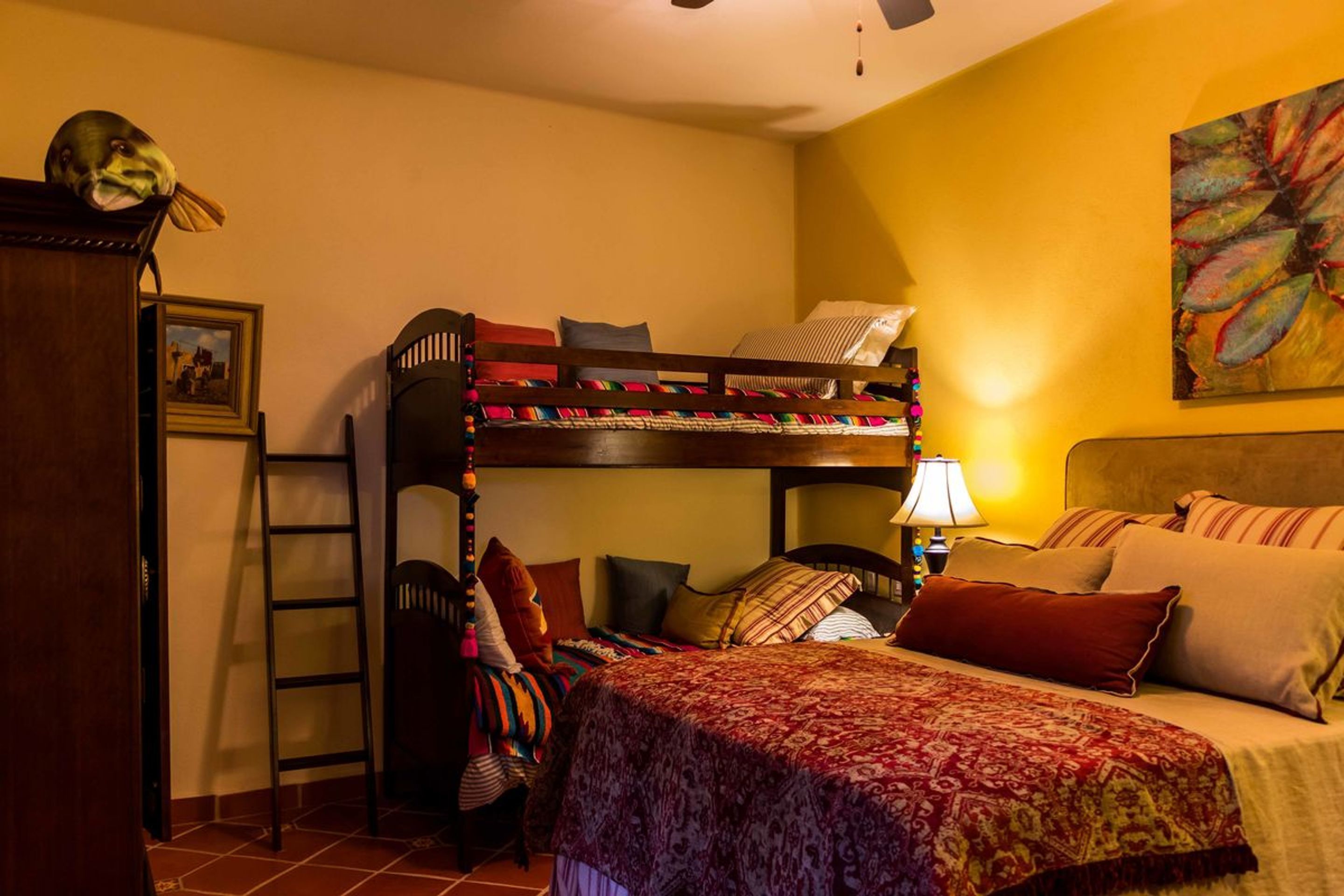 A dormitory room with bunk beds, a ladder, a bed with a patterned blanket, a nightstand with a lamp, and a painting on the wall.