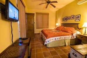 Una habitación de hotel con una cama, televisión, ventilador de techo, armario de madera y una mesita de noche con lámpara.