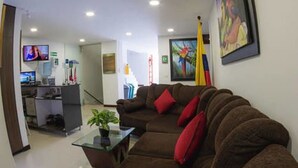 A living room with a brown sofa, a glass coffee table, and a potted plant. There is a television on the left side of the room.