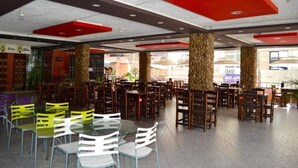 A spacious indoor dining area with multiple tables and chairs, featuring a modern ceiling design with red accents and natural stone pillars.