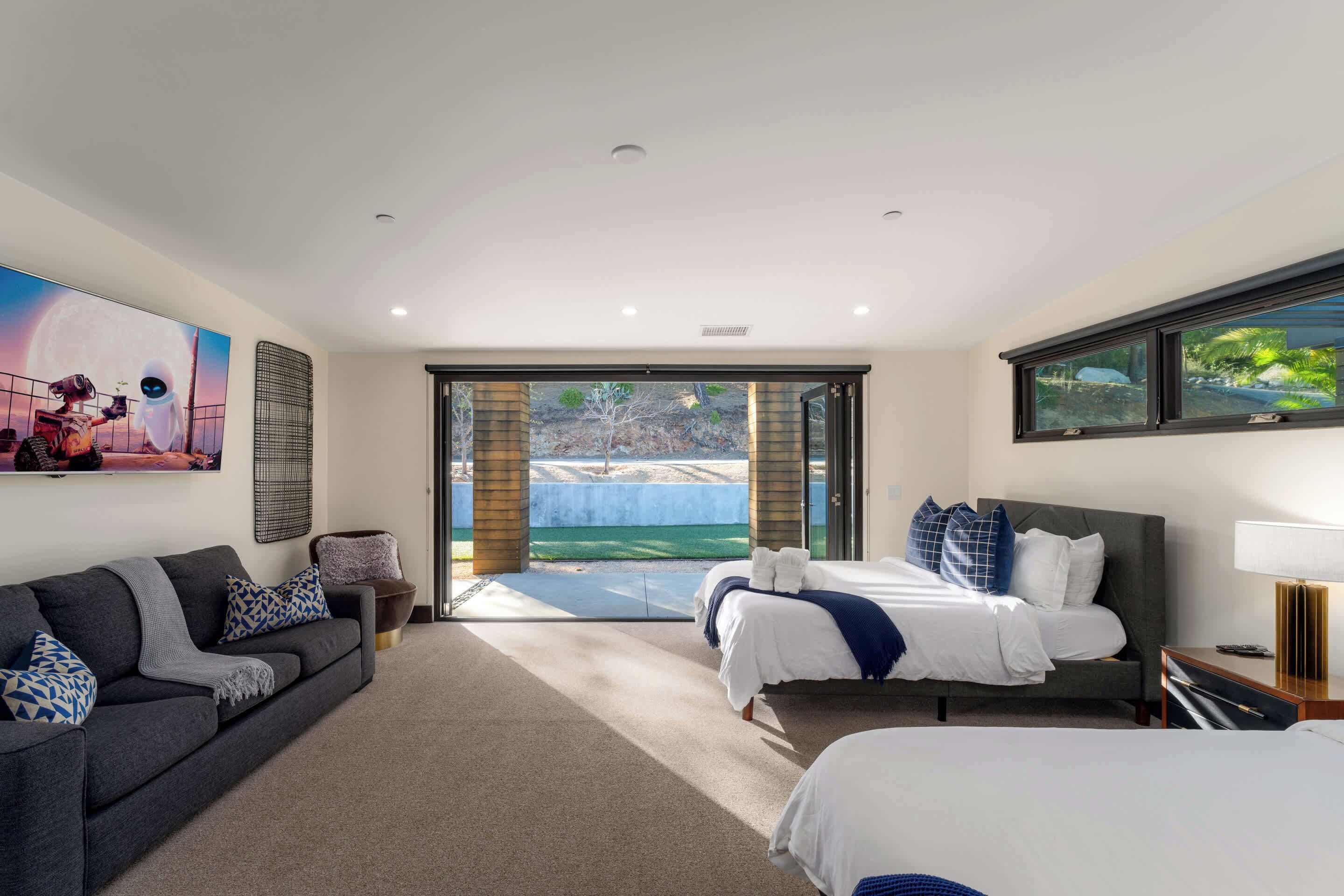 A modern hotel room with a large bed, a sofa, and a view of a landscaped area.