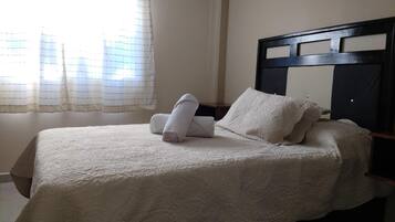 A neatly made bed with a patterned bedspread, a headboard with a geometric design, and a window with sheer curtains.