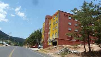 Um hotel com vários andares, fachada amarela e vermelha, entrada visível e uma estrada em primeiro plano.