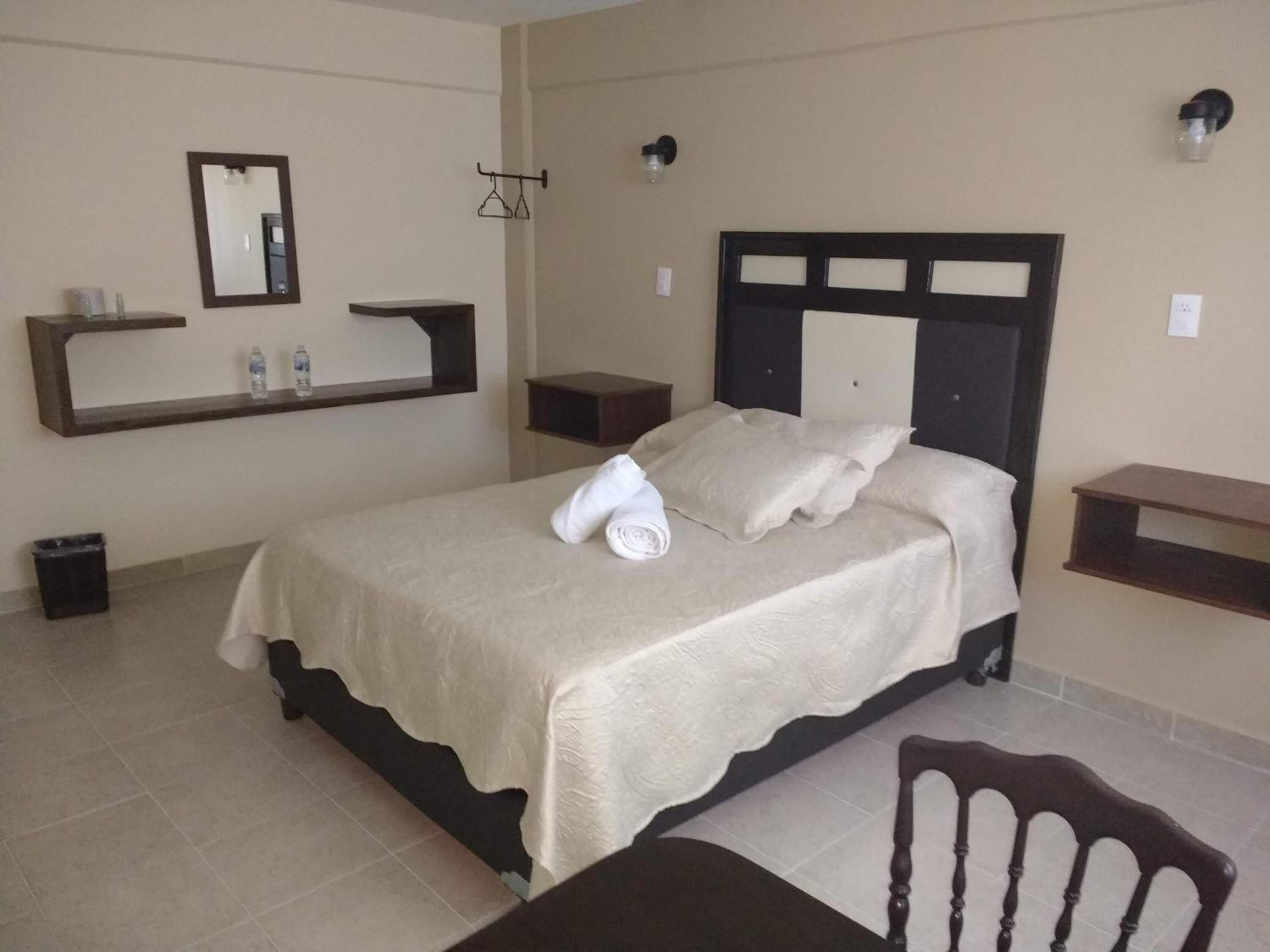 A neatly made bed with a headboard, a bedside table, a small shelf with toiletries, a mirror, and a wall-mounted light fixture.