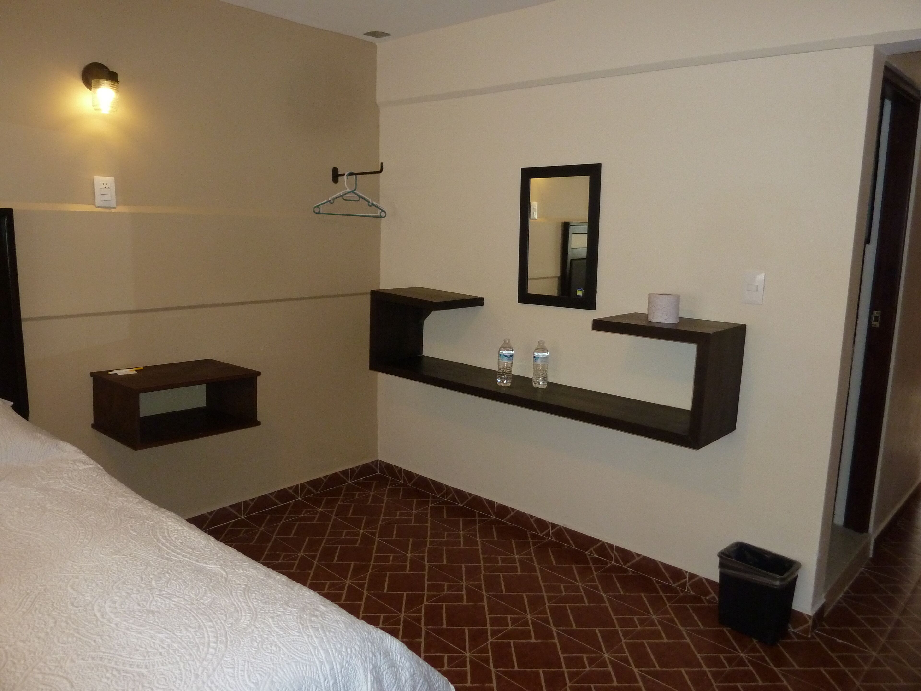 A hotel room with a bed, a small bedside table, a wall-mounted shelf with toiletries, a hanging clothes rack, and a trash bin.