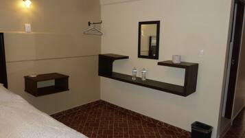 A hotel room with a bed, a small bedside table, a wall-mounted shelf with toiletries, a hanging clothes rack, and a trash bin.