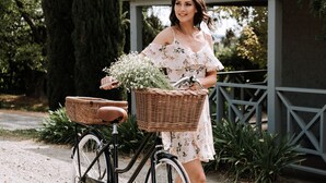 Une femme se tient à côté d’un vélo muni d’un panier en osier contenant des fleurs.