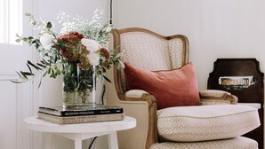 Une pièce dans laquelle on aperçoit une table ronde blanche, sur laquelle sont posés un vase de fleurs et une pile de livres. Un fauteuil à motifs, agrémenté d’un coussin rouge, est également visible.