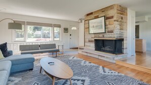 A modern living room with a stone fireplace, a blue sofa, a wooden coffee table, and a large window with blinds. - Private Beach • Hot Tub • Sauna • Game Room •  No-Wake Cove + Party Cove Access (Coloma)