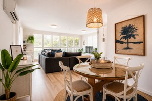 Ein modernes Wohnzimmer mit einem Glastisch, weißen Stühlen, einem dunklen Sofa und einem großen Fenster mit Jalousien.