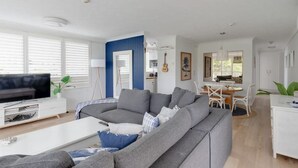 Een moderne woonkamer met een grijze hoekbank, een flatscreen televisie en een eethoek met witte meubels.