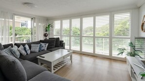 Een moderne woonkamer met een grijze bank, een witte salontafel en grote ramen met zonwering.