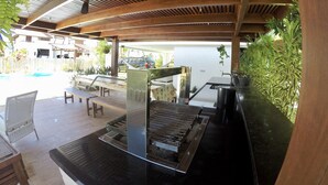 Een terras buiten met een grillplaats, zithoek en uitzicht op een zwembad.