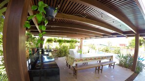 Een terras buiten met een houten pergola, een zwarte plantenbak, een tafel met banken en een zwembad op de achtergrond.