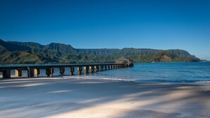 A beach with a wooden pier extending into calm waters, a small shelter at the end, and a mountain range in the background. - Spacious condo with Central AC, beautiful pool and relaxing hot tub (Princeville)