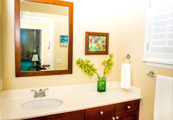 A bathroom with a large mirror, a sink, a framed picture, and a vase with flowers. - Spacious condo with Central AC, beautiful pool and relaxing hot tub (Princeville)