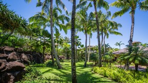 A tropical garden with palm trees, a stone wall, and a building with a red roof. - Spacious condo with Central AC, beautiful pool and relaxing hot tub (Princeville)