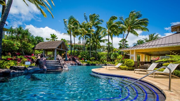 A swimming pool with a waterfall feature, surrounded by tropical plants and palm trees. - Spacious condo with Central AC, beautiful pool and relaxing hot tub (Princeville)