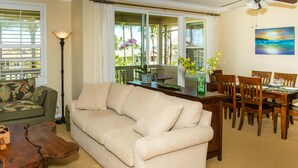 A living room with a beige sofa, wooden coffee table, and dining area with wooden chairs and a table. - Spacious condo with Central AC, beautiful pool and relaxing hot tub (Princeville)