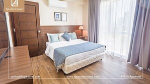 A spacious master bedroom with a large bed, a wooden wardrobe, a bedside table with a lamp, and a view of the outdoors through sheer curtains.
