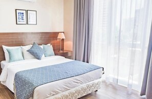 Un espacioso dormitorio principal con una cama grande, un armario de madera, una mesita de noche con lámpara y vistas al exterior a través de cortinas de gasa.