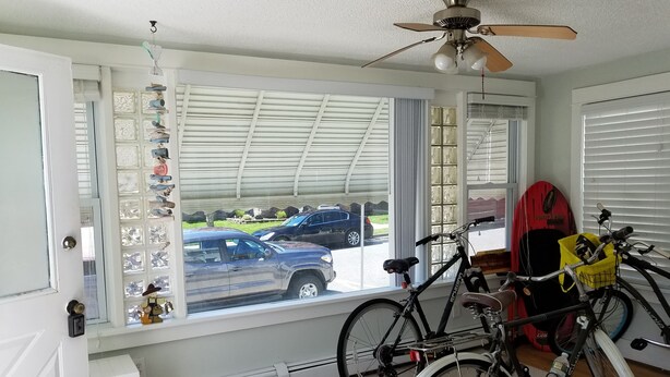 Een kamer met een groot raam dat uitkijkt op een parkeerplaats, een plafondventilator en een fiets met een mandje.