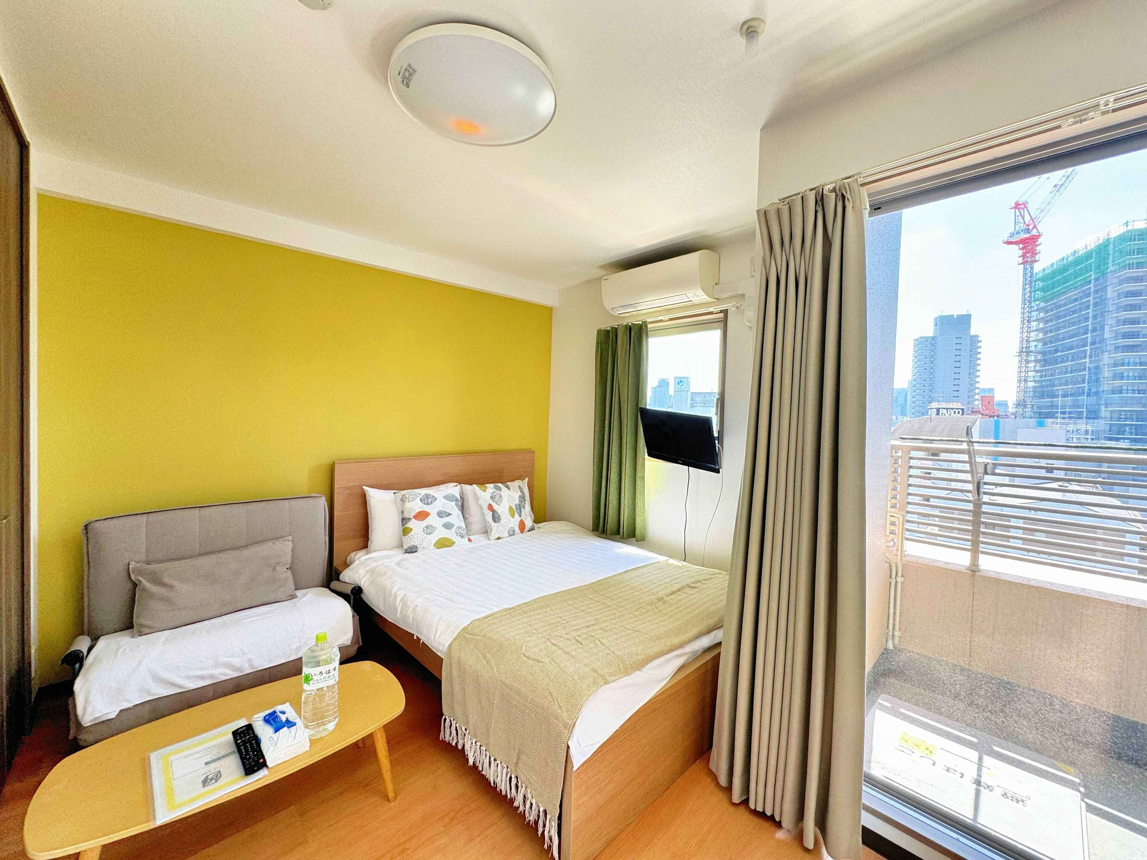 Quarto de hotel com cama, sofá, mesinha com garrafa e controle remoto, janela com cortinas e vista para a cidade.