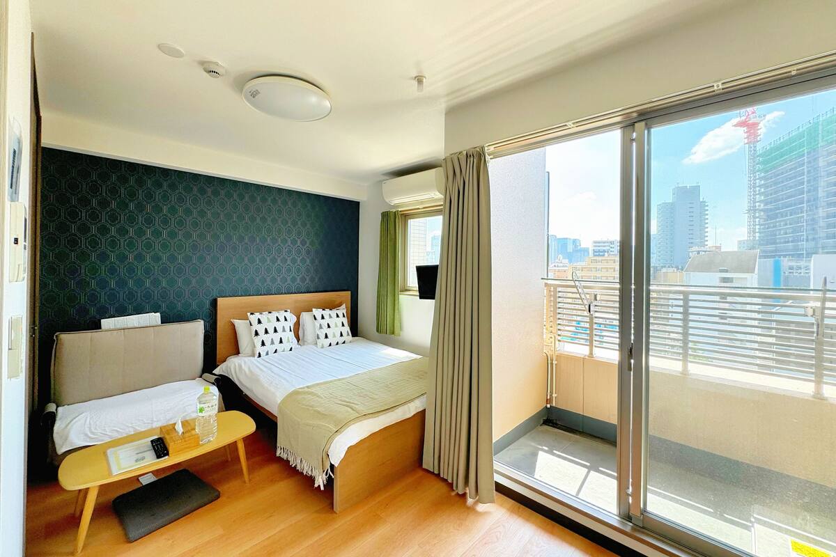 A hotel room with a bed, a small table with a chair, a window with a view of the city, and a balcony with a railing.