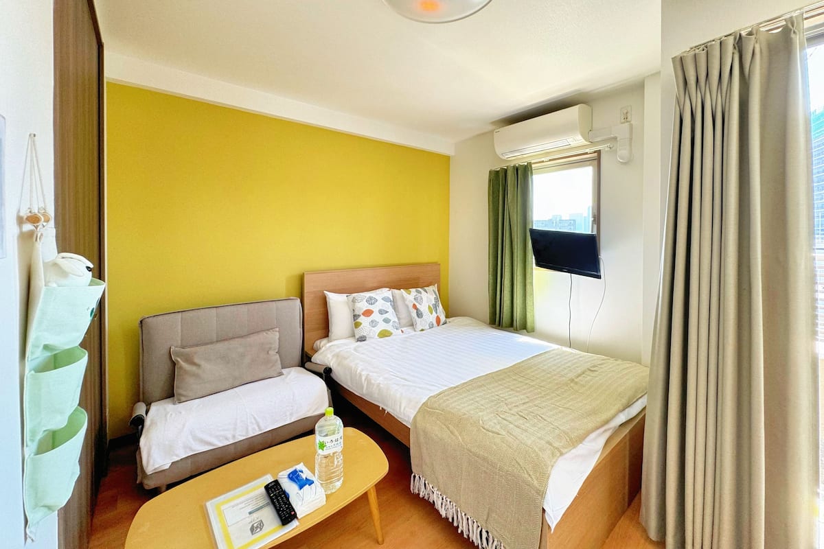 A hotel room with a bed, a sofa, a small table with a bottle and remote control, and a window with curtains.